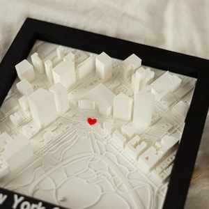 May include: A framed 3D map of a city, featuring white buildings and streets, with a small red heart marking a location. The map is set within a black frame, creating a striking contrast. The overall design is clean and modern.