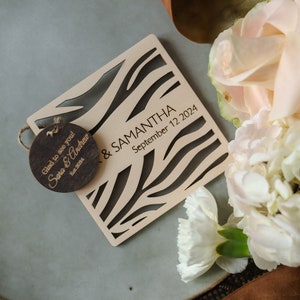 以下が含まれることがあります： ゼブラ柄のウッドウェディングギフト。ギフトには「Sara & Samantha」と「September 12 2024」の文字が刻まれています。小さな木製タグには「Glad to see you! Sara & Andrew」の文字が書かれており、ギフトに付けられています。