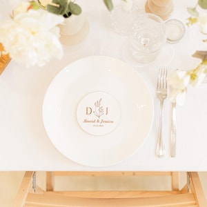 Puede incluir: Un posavasos redondo blanco con un dise&ntilde;o floral y las iniciales "D.J." en letras doradas. El texto "David & Jessica" y la fecha "19.11.2024" tambi&eacute;n est&aacute;n en letras doradas.