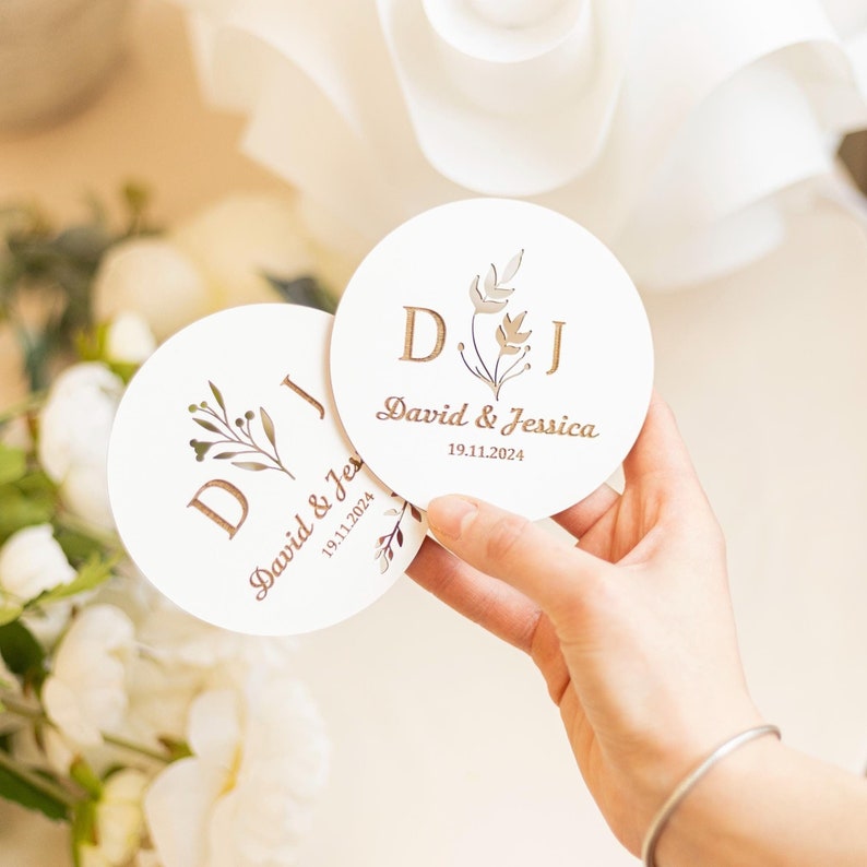 Puede incluir: Dos posavasos redondos blancos con un dise&ntilde;o floral y las iniciales "D.J." en dorado. El texto "David & Jessica" y la fecha "19.11.2024" tambi&eacute;n est&aacute;n en dorado.