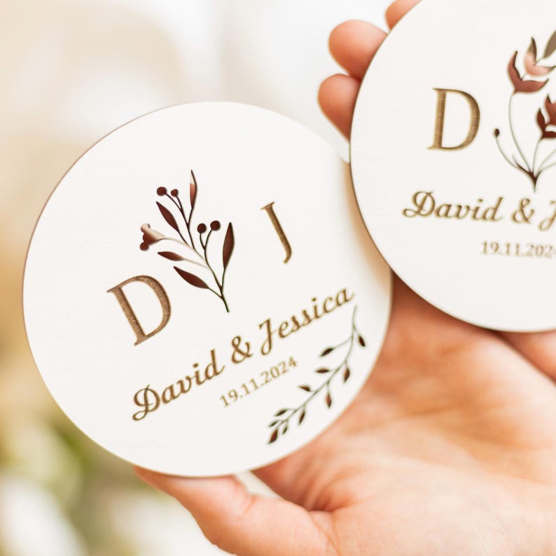 Puede incluir: Dos posavasos redondos de madera con un dise&ntilde;o floral y las iniciales "DJ" en dorado. Los posavasos est&aacute;n personalizados con los nombres "David & Jessica" y la fecha "19.11.2024".
