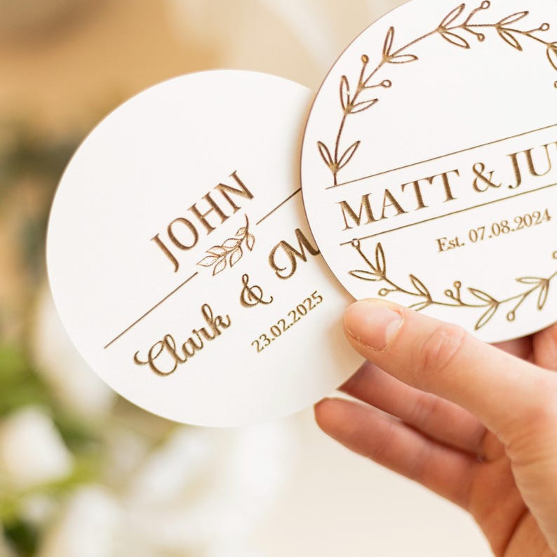 Puede incluir: Dos posavasos redondos de madera con texto grabado y dise&ntilde;os florales. El primer posavasos dice "JOHN Clark & M 23.02.2025". El segundo posavasos dice "MATT & JUL Est. 07.08.2024".