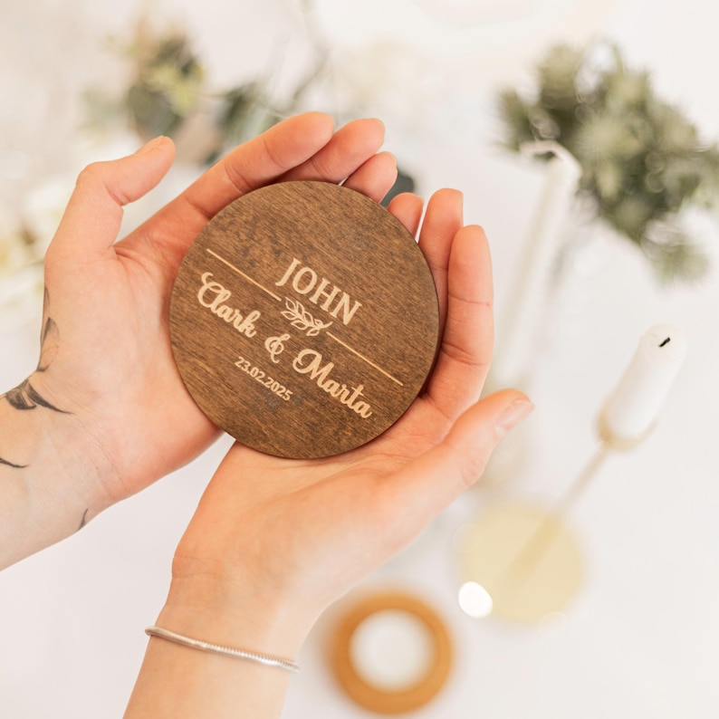 Puede incluir: Un posavasos redondo de madera con el texto "JOHN Clark & Marta 23.02.2025" grabado en oro.