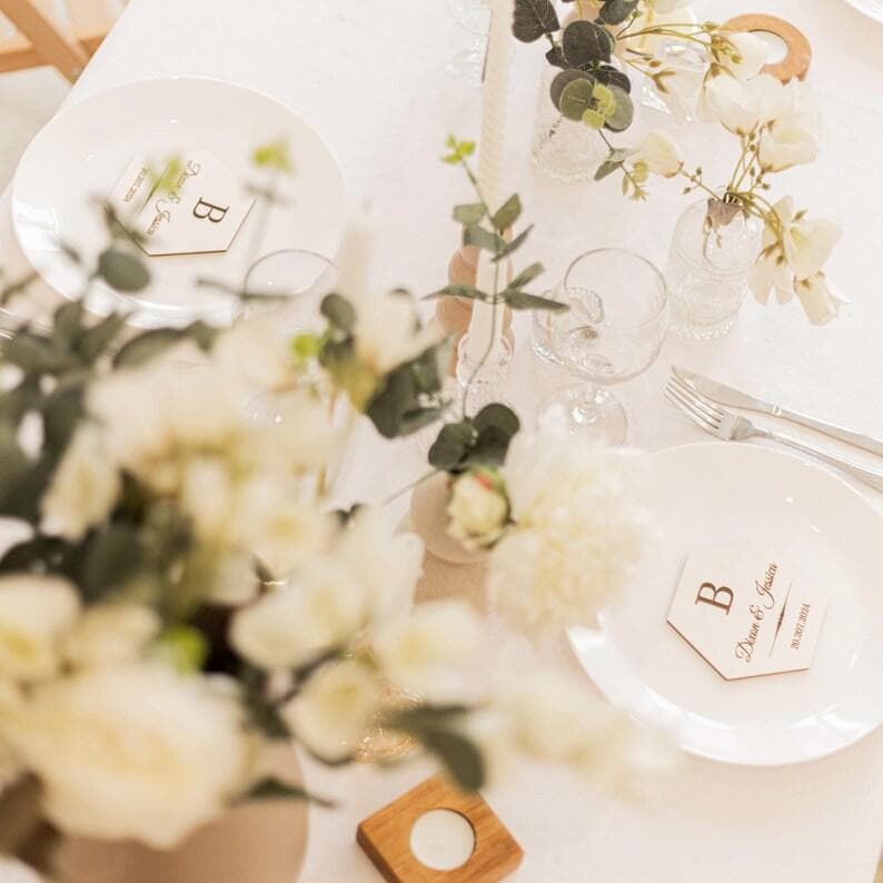 Puede incluir: Una mesa blanca con flores blancas, un plato blanco y una tarjeta de lugar de madera con la letra "B" y el texto "Diana & Jason" y la fecha "01.12.2018".