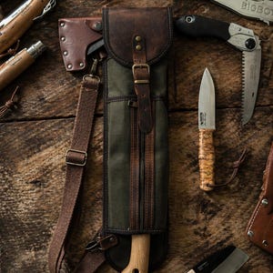 Op de afbeelding: Een assortiment buitentools, waaronder messen, een kleine bijl en een vouwzaag, is gerangschikt op een houten oppervlak. Een groene en bruine canvas en leren schede bevat een kleine bijl. De tools hebben houten en metalen handvatten.