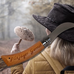 May include: A wooden mushroom knife with a curved blade and brush, engraved with "Pilzj&auml;ger Gernot". The knife is held near a black hat and a mushroom in a forest setting. The knife is a tool for mushroom hunters.