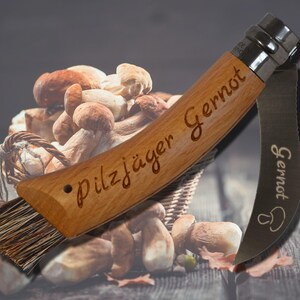 May include: A wooden mushroom knife with the text "Pilzj&auml;ger Gernot" engraved on the handle. The knife has a brush and a curved blade. The background shows a basket of mushrooms.