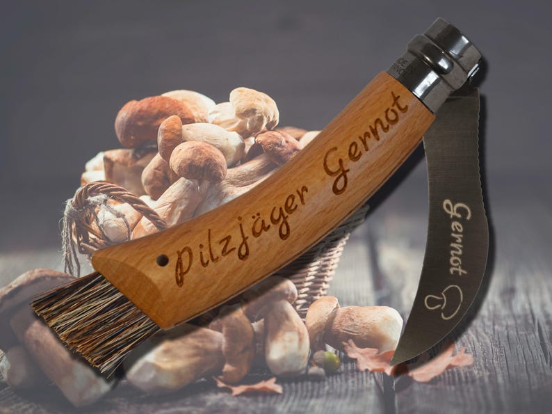 May include: A wooden mushroom knife with the text "Pilzj&auml;ger Gernot" engraved on the handle. The knife has a brush and a curved blade. The background shows a basket of mushrooms.