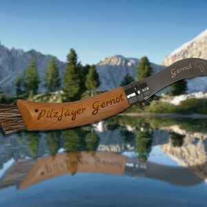 May include: A wooden-handled mushroom knife with a curved blade and a brush. The handle is engraved with "Pilzj&auml;ger Gernot" and the blade with "Gernot" and a mushroom illustration. The knife is designed for foraging.