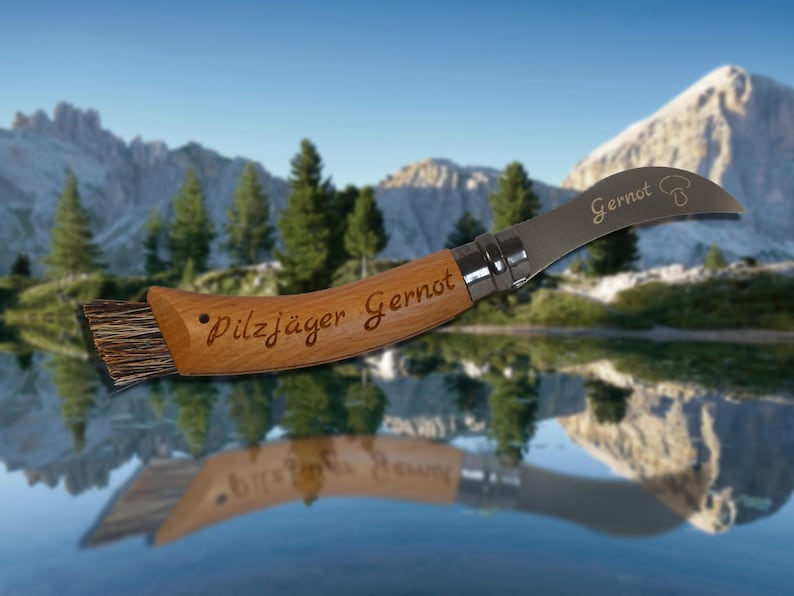 May include: A wooden-handled mushroom knife with a curved blade and a brush. The handle is engraved with "Pilzj&auml;ger Gernot" and the blade with "Gernot" and a mushroom illustration. The knife is designed for foraging.