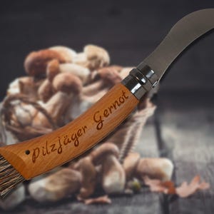 May include: A mushroom knife with a curved stainless steel blade and a wooden handle. The handle is engraved with the text "Pilzj&auml;ger Gernot". A brush is attached to the end of the handle. A basket of mushrooms is in the background.