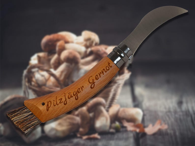May include: A mushroom knife with a curved stainless steel blade and a wooden handle. The handle is engraved with the text "Pilzj&auml;ger Gernot". A brush is attached to the end of the handle. A basket of mushrooms is in the background.