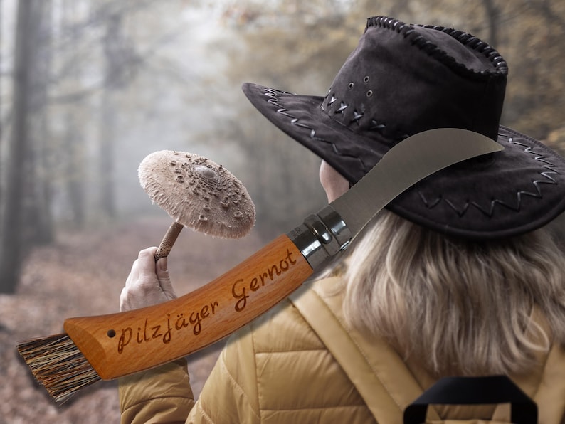 May include: A wooden mushroom knife with a curved blade and brush, engraved with "Pilzj&auml;ger Gernot". The knife is held near a black hat and a mushroom in a forest setting. The knife is a tool for mushroom hunters.
