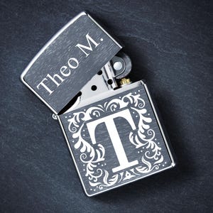 Könnte beinhalten: Ein silbernes Feuerzeug mit offenem Deckel, das den Text "Theo M." im Inneren zeigt. Auf der Vorderseite des Feuerzeugs befindet sich ein großer weißer Buchstabe "T", umgeben von einem verzierten floralen Design.