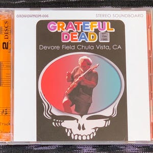 May include: A CD case with a colorful Grateful Dead logo and the text "Grateful Dead" and "Devore Field Chula Vista, CA". The case is orange and has the text "2 DISCS" on the side.