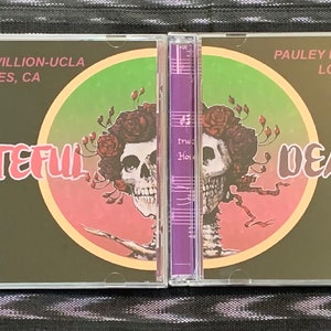May include: Two compact discs with a green, pink, and yellow skull graphic. The text "GRATEFUL DEAD" is printed on the discs. The text "PAULEY PAVILLION-UCLA LOS ANGELES, CA 11/25/79" is printed on the top of each disc.