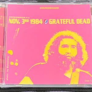 May include: A pink CD case with a yellow image of Jerry Garcia from the Grateful Dead. The text on the case reads "Soundboard", "Oxowowmom 514", "Nov. 3rd 1984 Grateful Dead", "photo: Jay Blakesberg", and "Disc 1".