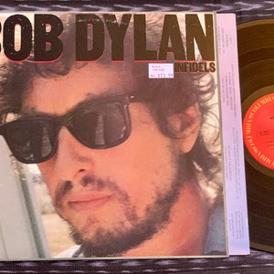 以下が含まれることがあります： ボブ・ディランのアルバム「インフィデルズ」のジャケットのアップ。ジャケットには、サングラスとひげを生やしたディランのモノクロ写真が写っている。アルバムタイトルはジャケットに金文字で印刷されている。