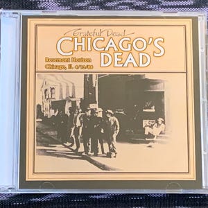 May include: A Grateful Dead CD titled Chicago's Dead, featuring a sepia-toned cover with a black and white photograph of people on a street. The text reads Grateful Dead, Chicago's Dead, Rosemont Horizon, and Chicago, IL 4/13/88 in a clear jewel case.