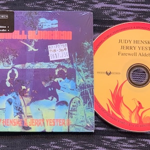 以下が含まれることがあります： 黄色い背景と赤いフェニックスのデザインが施されたCDケースとCD。CDケースには、ベンチに座っている2人の写真が写っている。CDケースのテキストには「Farewell Aldebaran」と「Judy Henske & Jerry Yester」と書かれている。CDのテキストには「Judy Henske & Jerry Yester Farewell Aldebaran」と書かれている。