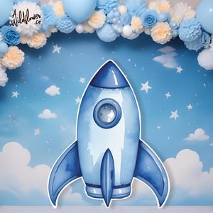 Op de afbeelding: Een blauwe en witte waterverf raket-uitsparing met een raam. De raket is tegen een blauwe achtergrond met witte wolken en sterren.