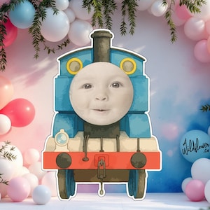 Könnte beinhalten: Ein blauer und orangefarbener Zeichentrickzug mit einem Babygesicht in der Mitte, umgeben von rosa, weißen und blauen Luftballons. Der Zug hat einen schwarzen Schornstein und eine rote Front. Der Hintergrund ist ein sanfter rosa-blauer Farbverlauf.