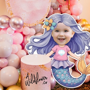 May include: A pink and gold backdrop with a balloon arch and a mermaid cutout with a girl's face. The mermaid has purple hair, a pink shirt with a seashell, a purple skirt, and a blue and green tail. The text "A Wildflower Co." is on a pink and gold cylinder.