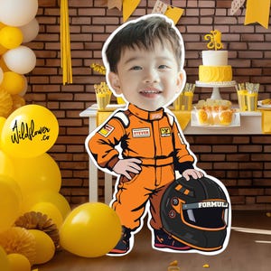 May include: A cartoon cutout of a child in an orange racing suit holding a black helmet with the word "FORMULA" on it. The background includes a brick wall, yellow balloons, and a table with a cake and cupcakes.