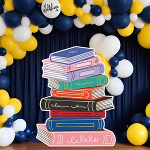 Puede incluir: Una pila de libros coloridos e ilustrados se alza frente a una cortina azul marino y un arco de globos. Los globos son blancos, amarillos y azul marino. Los libros son de varios colores, incluyendo rosa, azul y verde. El texto "Wildflower .co" está en un globo blanco.