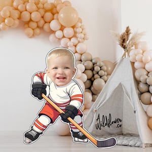 Puede incluir: Ilustración de dibujos animados de un bebé sonriente con uniforme de hockey, sosteniendo un palo de hockey. El bebé lleva una camiseta blanca con rayas rojas, pantalones cortos rojos, guantes negros y patines negros. Una tienda tipi y un arco de globos en el fondo.