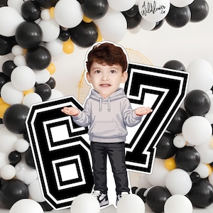 Puede incluir: Un recorte de dibujos animados de un niño pequeño con una sudadera con capucha gris y pantalones negros, de pie frente a un gran número 67 en blanco y negro. El fondo presenta un arco de globos en negro, blanco y amarillo. También es visible un globo con el texto "Wildflower.co".