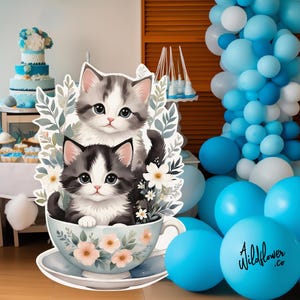 May include: Two adorable kittens, one gray and white and one black and white, are sitting in a blue and white teacup with floral designs. The kittens are surrounded by green leaves and white flowers. The teacup is on a white saucer with a floral design. The background is a blue and white balloon arch.