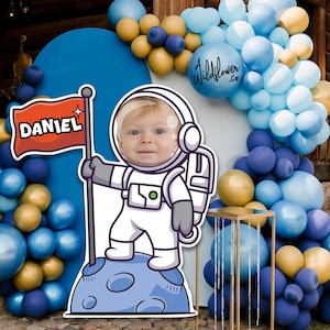 Op de afbeelding: Een feestdecoratie met een kindergezicht in een astronautenpak, met een vlag met de naam "DANIEL". De achtergrond bevat blauwe, gouden en witte ballonnen, wat een feestelijke sfeer creëert.