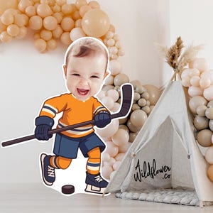 Peut inclure: Illustration de dessin animé d'un bébé en uniforme de hockey, tenant une crosse et un palet. Le bébé porte un maillot orange, un short bleu et des patins noirs. Un tipi gris avec le texte "Wildflower.co" est en arrière-plan, avec une arche de ballons.