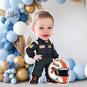 以下が含まれることがあります： 黒いレーシングスーツを着た幼い子供の漫画の切り抜きで、レーシングヘルメットを持っています。ヘルメットは白地に赤とオレンジのストライプで「FORMULA」の文字が入っています。背景には、青、白、金色の風船のアーチがあります。