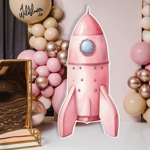 Op de afbeelding: Een roze raketdecoratie met een rond raam en vinnen, omringd door roze, gouden en crèmekleurige ballonnen. Een groot gouden cijfer één staat op de voorgrond en een ballon met de tekst "Wildflower .Co" is zichtbaar.