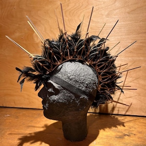 Puede incluir: Una corona decorativa con base de plumas negras, detalles metálicos dorados y largas y finas púas. La corona está sujeta a una banda elástica negra y se exhibe sobre una cabeza de maniquí negra.