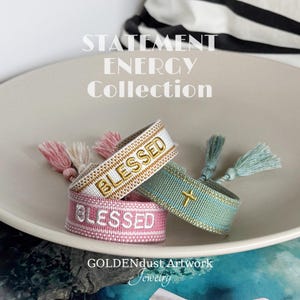 Könnte beinhalten: Drei bestickte Armbänder mit dem Wort "BLESSED" und einem goldenen Kreuz. Die Armbänder sind weiß, rosa und türkis, mit passenden Quasten. Der Text "STATEMENT ENERGY COLLECTION" und "GOLDENDUST Artwork Jewelry" ist ebenfalls sichtbar.