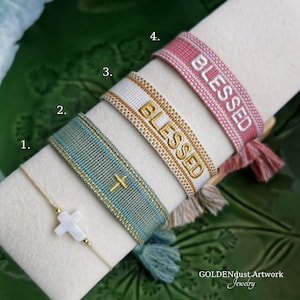 Könnte beinhalten: Gezeigt werden vier Armbänder. Eines ist ein zartes Armband mit Kreuzanhänger. Die anderen sind gewebt, mit dem Wort "BLESSED" auf zwei gestickt. Das vierte Armband ist rosa mit weißem Text. Die Armbänder haben Quasten.