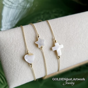 Könnte beinhalten: Drei goldene Kettenarmbänder mit weißen Muschelanhängern. Die Anhänger sind ein Herz, ein Stern und ein Kreuz.