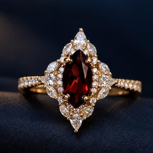 May include: A gold ring featuring a central marquise-cut deep red garnet gemstone. The stone is surrounded by a halo of small marquise-cut and round white stones. The ring band is thin and decorated with a row of small round white stones.