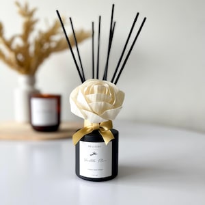Könnte beinhalten: Ein schwarzer Schilf-Diffusor mit einer cremefarbenen rosenförmigen Oberseite und schwarzen Schilf. Die Flasche hat ein goldenes Band und ein Etikett mit der Aufschrift "DS LUXURY Vanilla Flower Luxury Reed Diffuser". Ein dekorativer Raumduftartikel.