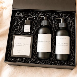 Puede incluir: Una caja de regalo negra que contiene un jabón de leche de cabra, una crema de manos y cuerpo, un gel de lavado y un difusor para coche Cedar & Amber. Cada producto tiene una etiqueta blanca con texto negro sobre un lecho de papel triturado.