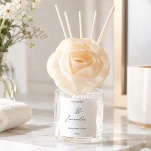 May include: A clear glass reed diffuser with a cream-colored rose-shaped top and white reeds. The label reads "Lavender Luxury Reed Diffuser" and the bottle contains 184 ml of liquid. The diffuser is on a marble surface.