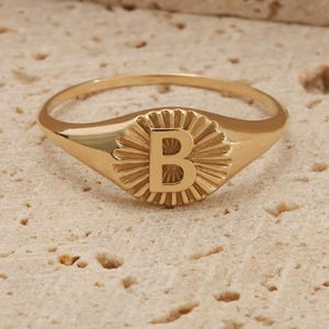May include: A gold signet ring with the letter "B" in a sunburst pattern. The ring is displayed on a textured surface. The ring's design and polished finish are highlighted. This piece of jewellery is a classic and personalised accessory.