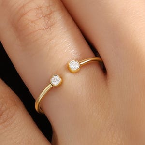 May include: A gold-colored open ring with two small, round, clear gemstones set at each end. The ring is shown on a person's finger, with the ring's design creating an open space between the gemstones.