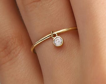 May include: A delicate gold ring with a small, dangling, round, clear gemstone. The ring is simple in design, with a thin band and a small charm. The ring is shown on a person's finger.