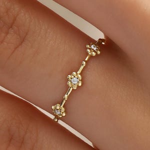 May include: A delicate gold ring featuring a series of small, flower-shaped designs. Each flower has a central, sparkling gemstone. The ring is shown on a person's finger, highlighting its elegant and minimalist style.