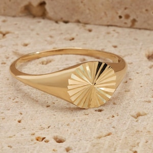 May include: A gold ring with a sunburst design. The ring features a smooth band and a circular, textured face. The ring is made of gold and is displayed on a light-coloured, textured surface.