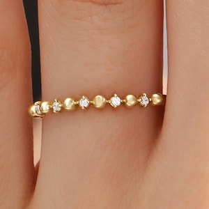 Op de afbeelding: Een delicate gouden ring met een reeks kleine, gelijkmatig verdeelde kralen en afgewisseld met kleine, heldere edelstenen. De ring wordt getoond op de vinger van een persoon, waarbij de fijne details en het elegante ontwerp worden benadrukt.